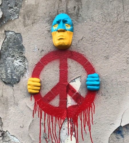 Vredesteken in rood op muur geschilderd. Een huilend gezicht en handen in blauw en geel, vormen een stuurman achter een stuur.