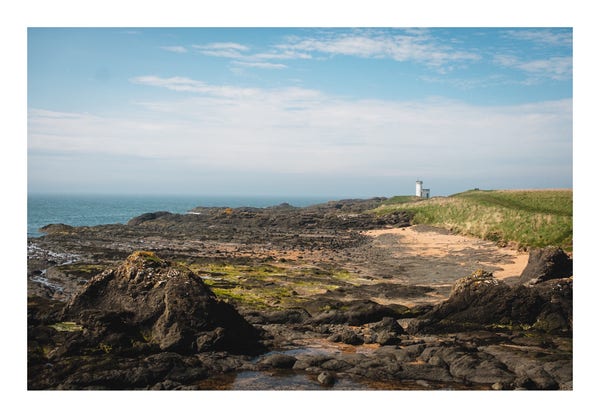 Rocky coastline with a lighthouse in the distance, blue sky, and calm sea.