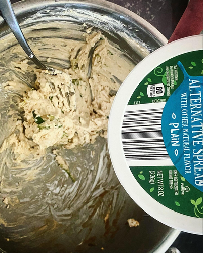 A steel mixing bowl with the creamcheese-like filling and a fork inside of it. The Aldi-brand vegan creamcheese container is in the frame on the right half of the picture, showing the OU hechsher. It’s a round tub with a dark green background and lighter green cartoon leaves in lighter shades. A dusty cerulean bubble with white writing says “alternative spread with other natural flavor plain” with a barcode underneath it that takes up about a sixth of the lid. 
