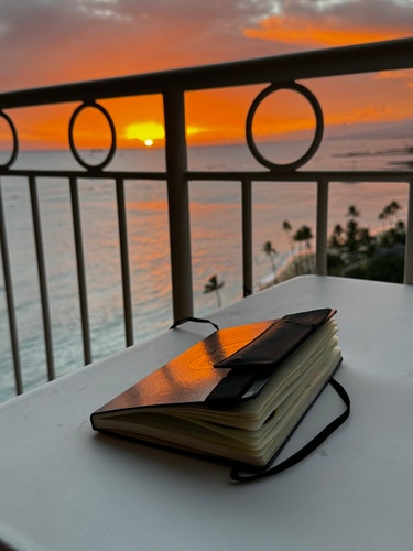 A closed notebook rests on a table with the sunset reflecting on its cover, against a backdrop of a scenic sunset over water viewed from a balcony.