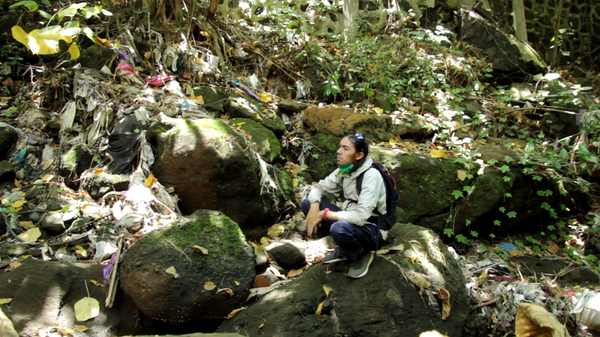A man crouches in the middle of a rocky river bank. He is surrounded by moss and trash.