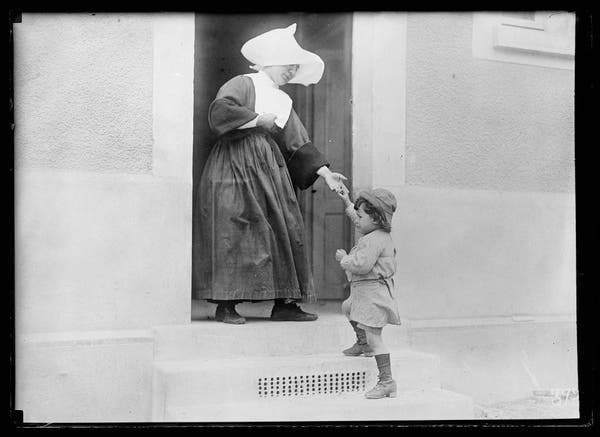  The image is a vintage black and white photograph. It captures a tender moment between two individuals at an old-fashioned doorway. On the left, there's a woman who appears to be from a religious or charitable organization, possibly a nun or a member of the Sisters of St. Vincent de Paul given her attire. She is holding what looks like a white cloth in her hand, which could be part of her work or an offering. On the right, there's a child, perhaps a toddler or young boy, looking up at the woman with a curious or expectant expression. The setting appears to be a residential building, and the architecture suggests it might be from a bygone era. There is no text visible in the image to provide additional context.