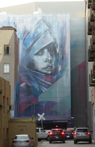 Streetartwall. A beautifully designed mural of the singer Lady Gaga was sprayed/painted on the exterior wall of a multi-story modern building. The background is gray/blue. It shows a woman wearing a metal knight's helmet with an open visor. She is looking in the direction of the viewer. The mural is sprayed entirely in shades of gray and blue and appears blurred, intangible. A few dark red splashes of color enliven it. (In the photo, a road between high-rise buildings leads directly to the mural. Some cars with red brake lights are driving towards the house)
Info: This was a tribute to Lady Gaga who played the Golden One Center on the night this wall was finished (on the back of the Golden One Center)