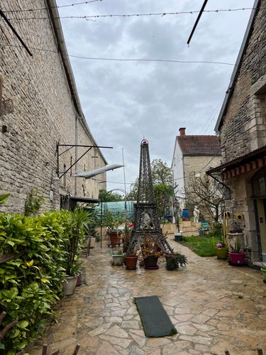 The Eiffel Tower at Nuits s/ Armançon. It’s in a stone paved yard with a barn to the left and a house to the right. Bushes line the yard.