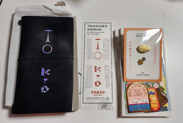 A black leather traveler's notebook. It has the word Tokyo spelled out on the front in a holographic purple using landmarks and culturally relevant items such as sakura blossoms and sushi.

Beside it is the slip with the tokyo design again and Japanese text. There are two brass charms, and taiyaki fish and a sakura blossom. Under the packet are some Japanese themed stickers.