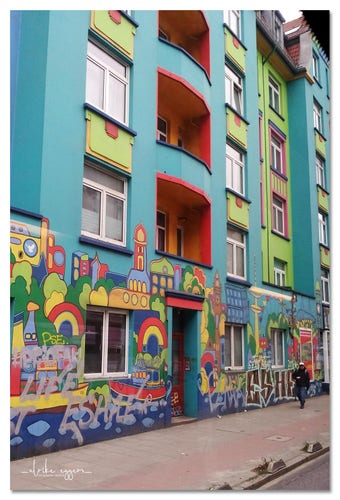Ein 6-stöckiger Altbau, durch einen nicht sehr breiten Fuß- und Radweg von der Straße getrennt. Die Hausmauern sind abwechselnd türkis-blau und hellgrün gestrichen, die Wände der nach innen gebauten Balkone sind gelb und orange. Auf der ganzen Höhe des Erdgeschosses und über die gesamte Breite des Hauses ist ein farbenfrohes Wandbild, das in naiver Malerei die Stadt Hamburg mit diversen Sehenswürdigkeiten wie der Elbphilharmonie, den Landungsbrücken und der Elbe mit Schiffen darauf darstellt.