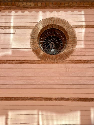 Round window on the side of a chapel in Murcia, Spain. The building is made of light tan brick, and the window is barred. The light reflected off the more modern opposite, unseen building, create a play of light on the wall. 