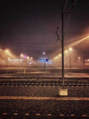 On a foggy railway station the camera looks from one platform towards the other Platforms with the tracks running left to right. On the next platform the sign shows “Leipzig Hbf”. The platform lights cast light rays through the fog, which makes the fog glow yellowish.