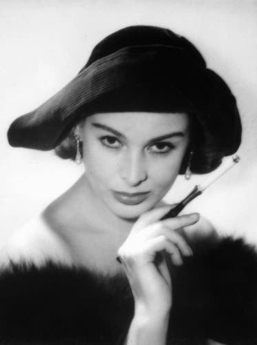  The image is a black and white photograph featuring a woman in mid-century fashion. She has a large, wide-brimmed hat that drapes over her hair. Her eyes are looking directly ahead, conveying a sense of confidence or intrigue. She has a cigarette held up to her mouth, suggesting a pose often associated with smoking or an air of sophistication during the 1950s era. There's a fur stole draped around her shoulders, adding to her elegant appearance. Her lips are parted slightly, and she has a slight smirk on her face that hints at a playful or enigmatic personality. The style of the photograph is reminiscent of classic Hollywood portraits from the 1950s era.