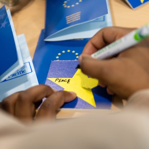 A photo of a visitor writing the word "Peace" on a piece of yellow paper. There are books with the EU logo on the cover. 