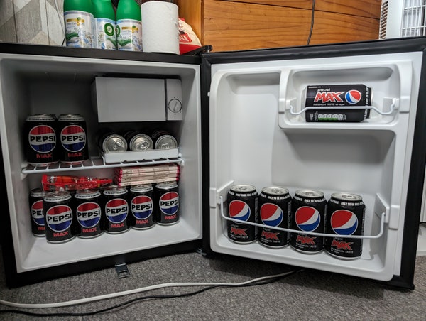 a mini fridge filled to the brim with pepsi max, with some galaxy cookie crumble bars and a bag of munchies as well