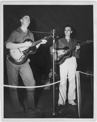  This is a black and white photograph featuring two men standing in front of microphones, performing music. The man on the left appears to be holding an acoustic guitar while playing, and he is wearing a striped shirt. On his right stands another man, also holding an acoustic guitar and wearing a similar style shirt with long sleeves. Both are looking towards the camera, engaging in performance. They are positioned in front of what seems to be a stage or concert setup.

The background suggests an outdoor setting, possibly a festival, as indicated by the text "Mountain Music Festival, Asheville, North Carolina." The style of clothing and the quality of the image indicate that it is likely from mid-20th century judging by the clothing and the quality of the image. 