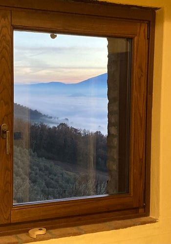 A wood framed window with a tarnished brass handle mounted into a yellow plastered wall. The window takes up most of the shot. Through the window, olive trees grow on a hill that slopes downward and more trees beyond. There is fog over the valley below and blue mountains rise in the background. It is sunset and the cloudy skies are just starting to color.