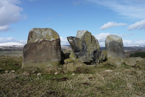 Recumbent and flanker stones at Ardlair. A  massive but partially shattered stone lying lengthways, flanked by two upright stones. The setting is an open grassy hilltop, with higher snow covered hills on the skyline behind. It's sunny and sky is blue with some white clouds.