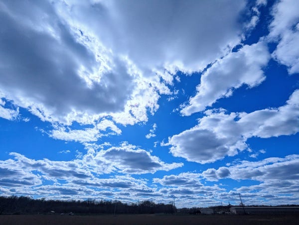 An empty field underneath the big blue sky with patchy clouds and sun