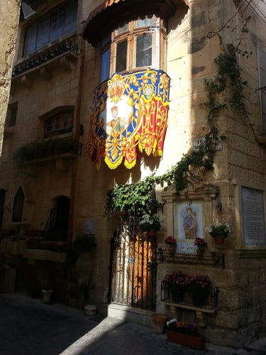 House on the corner of the Triq Id-Dejqa and the Triq Bir-Il-Ljun at Ir-Rabat on Malta, with a balcony cover and tiles featuring San Ġużepp or St Joseph. Photograph taken on the 21st of March 2014.