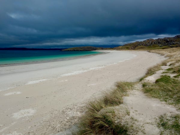 A white sand beach curves round, edged by dunes and Marram grass. The sea is bright turquoise near the shore, a darker blue further out. In the distance a rocky horizon and a really heavy stormy sky.