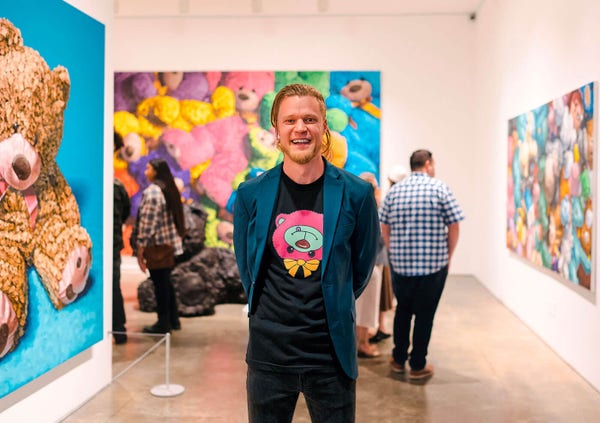 A photo showing artist Brent Estabrook smiling at the camera and standing next to some of his artworks in an art gallery.