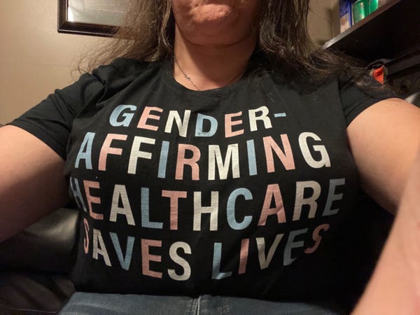 A photo of a woman’s chest; her t-shirt reads “Gender affirming care saves lives” in white, pink, and light blue letters.  