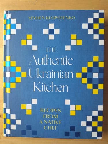 Front cover of The Authentic Ukrainian Kitchen by Yevhen Klopotenko.

The author's name is at the top in yellow.

The book title appears across the middle in white.

'Recipes from a native chef' at the bottom, in yellow.

The artwork is minimal and striking: a blue background, with a faint grid invoking the idea of square tiles. Symmetrical tiles patterns of white, dark blue and yellow, like a mosaic. Some of the dark blue tiles catch the light and it can be seen they have a metallic sheen.