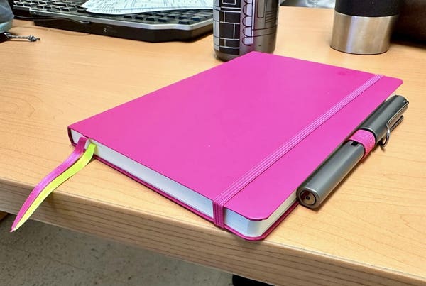 Closed pink notebook with elastic band and bookmark ribbon on a wooden desk, alongside a pen and a partial view of a keyboard and a metal water bottle.