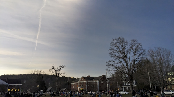 A picture of people gathered on a college green, staring upwards. Thin clouds in the sky are illuminated in half the sky, and dark in the other half.