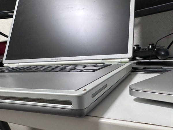 An open PowerBook G4 laptop on a desk with another closed laptop and peripherals nearby. The base of the G4 is thicker than the whole closed MacBook Pro 