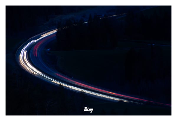 Long exposure photograph of a 90 degree road bend with heavy traffic through the forest at night, seen from ~150m above.