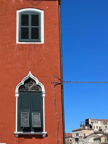 Colour photo, the left half of which is the edge of a rich terracotta-coloured building, in which are set two windows: the top one is small and squat and gently curved at the top, has a white marble surround, and dark green closed slatted shutters. The window beneath is taller and more elegant, topped by a beautiful Gothic arch, and the lower half of its dark green slatted shutters are angled outwards towards the viewer. The right half of the image is at the top clear intensely blue sky, at the bottom a distant view of higgledy-piggledy Venetian rooftops, and just under halfway up, a cable which has been run up the side of the building and then at a right angle off the edge into the void...and whatever it connects to on the other end is out of shot. 