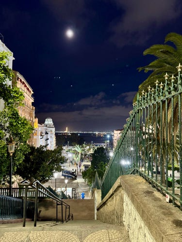 Nighttime Algiers city view from stairway with moon in the sky, palm trees, ornate railing, and distant lights along a waterfront promenade.

Sidi M‘Hamed, Alger, Algeris