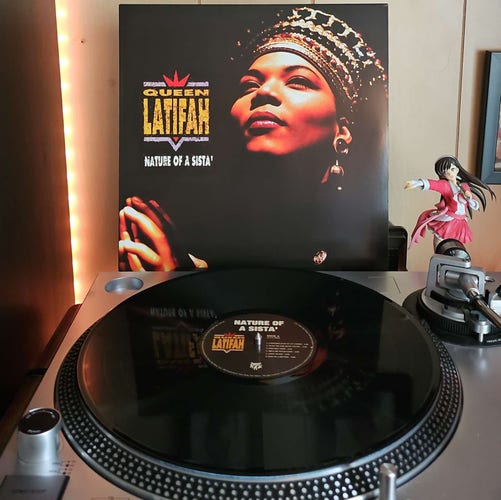 A vinyl record sits on a turntable. Behind the turntable, a vinyl album outer sleeve is displayed. The front cover shows Queen Latifah looking upward with her hands together. 