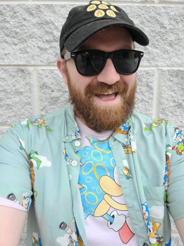 Selfie of a balding, bearded white man standing against a concrete block wall. He is wearing a black hat with Dragon Balls on it and a pink shirt with Sonic the Hedgehog in pastel colors with a green Hawaiian style casual button down shirt covered in various images of Sonic the Hedgehog and Tails. He is also wearing black sunglasses.
