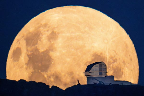 The yellow disc of the full moon dominates the image. The moon has nearly risen above the horizon runninb along the bottom of the image, but is just cut off from being a perfect circle at the bottom. Rubin Observatory is silhouetted toward the right side of the moon, shaped like a boot with low service building extending to the right and shiny angular dome sticking up. A small-looking crane sits to the left of the observatory.