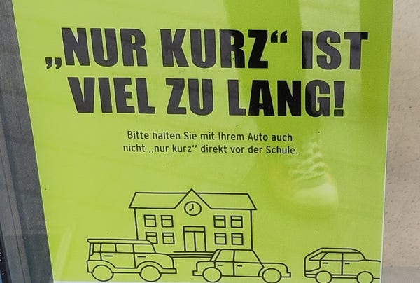 Plakat hinter einer Scheibe: "Nur Kurz" ist viel zu lang! Bitte halten Sie mit Ihrem Auto auch nicht nur kurz direkt vor der Schule.