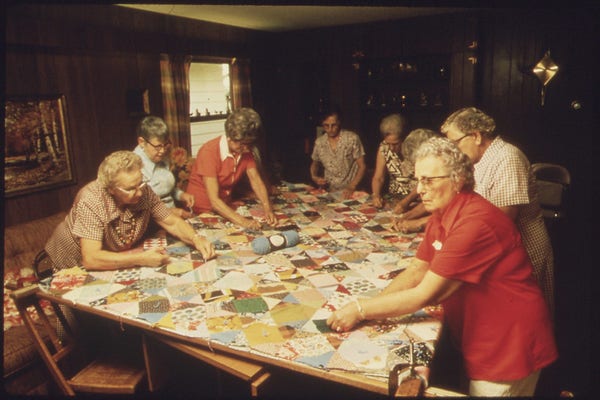 Eight woman mostly with grey hair and glasses work together on a quilt in a wood=paneled room