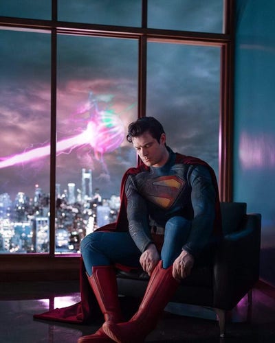 Official promotional image for the upcoming James Gunn Superman movie. Actor David Cornswet is sitting down in full Superman costume and perfect Superman spitcurl, pulling on a boot.  His right shoulder area looks like it has been burned or has some kind of ash coloring on it. 

In the background is a window, where a city (presumably Metropolis) glows slightly under thick dark clouds. Some kind of glowing sphere hovers above it, and a large beam of light shoots from the sphere onto the city below.