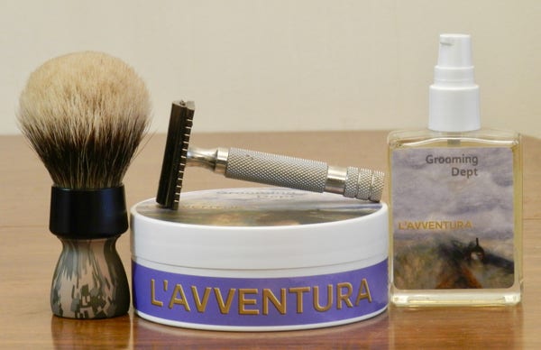 A shaving brush with a camo handle and a rounded silvertip badger knot stands snext to a tub os shaving soap with a bluish-violet label on the side with "L'Avventura" in gold caps. Lying on top is a DE razor that has a brown head and a stainless-steel bulldog handle. At the right in a small rectangular glass bottle with a white spray cap and a label that shows an Impressionistic country lain with a skill filled with white clouds. At the top appears "Grooming Dept" and across the middle, in gold, "L'Avventura."