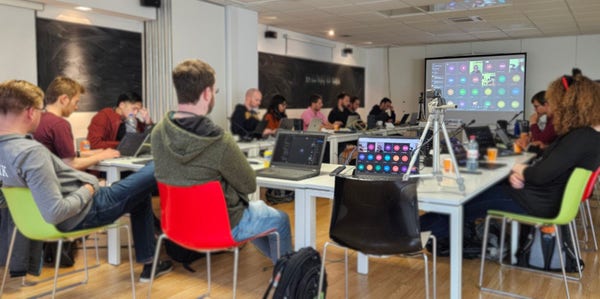 Several people discussing during the 2024 Display Next Hackfest in the Igalia Headquarters in Coruña, Spain. Tables and chairs are set up forming a U-shape with participants seating around it.