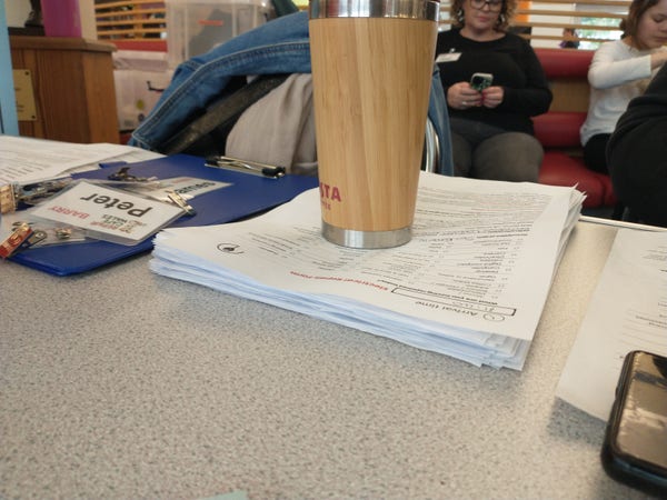 A thick stack of repair cafe forms on a table weighed down by reusable coffee cup.