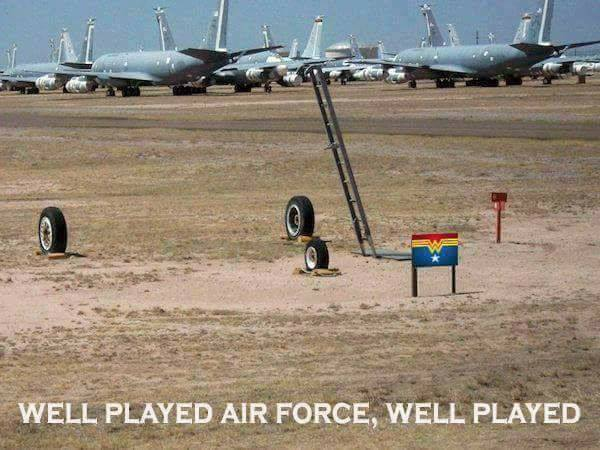 Picture of a US Air Force base with cargo planes parked in the background. In the foreground, in the empty sand, are three airplane wheels in a triangular configuration held in place by chocks, and an inclined ladder that seems to go to the empty air above the wheels. A small sign in front of this has the Wonder Woman logo, as if it's her reserved parking spot. The caption reads "Well played, Air Force. Well played."