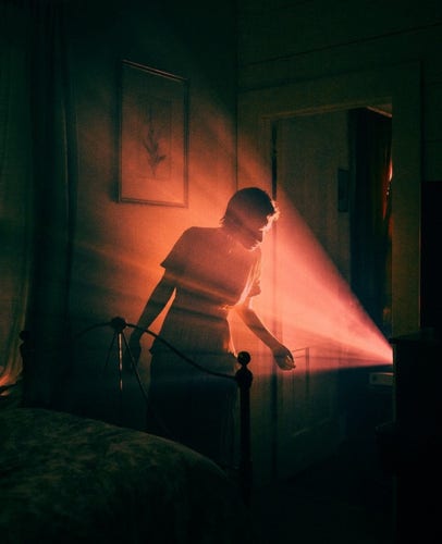 Photography. A mystical color photo of a young woman in front of a light source. The scene is a bedroom with pictures on the wall, a metal bed in the foreground and a young short-haired woman in a white dress behind it. The room is bathed in red light that appears to come from a tablet or television. The young woman leans forward and looks in.
Info: Model: Artist M.K.Sadler.