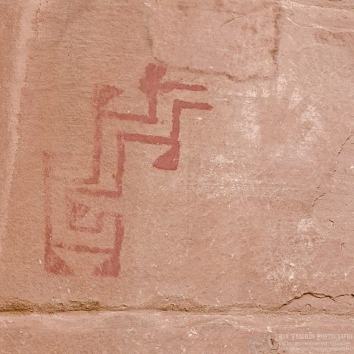 A color photo of an abstract design painted by a prehistoric Puebloan on a dark tan sandstone cliff face. The design in red colored and is a series of two parallel lines in a zigzag with little triangle like serifs on some of the corners. To the right of the design is a negative hand print made by holding ones hand against the stone wall and blowing paint that leaves and outline of the hand. It was made with a white color "paint." 