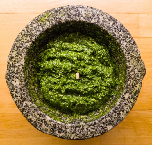 A black and white speckled marble pestle filled with vivid green wild garlic pesto, with a single pine nut in the middle. The background is a yellow wooden worktop.