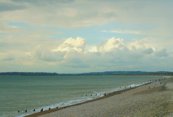 A stony beach, with wooden groynes, sloping down to a green sea. The beach is to the right, and the sky above is full of fluffy clouds. 