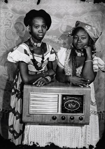 Black and white photo of two young Black women in patterned dresses posing with their arms over a large radio