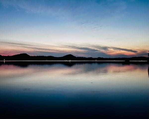 Twilight landscape showing a calm lake with cloud reflections which look like the feathered tail of an arrow, silhouetted hills on the horizon, and a colorful sky.