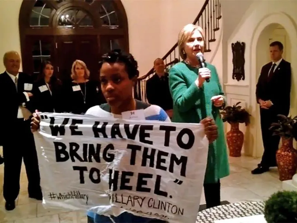 A young black woman holding up a sign including a quote from Hillary Clinton: "We Have To Bring Them to Heel".