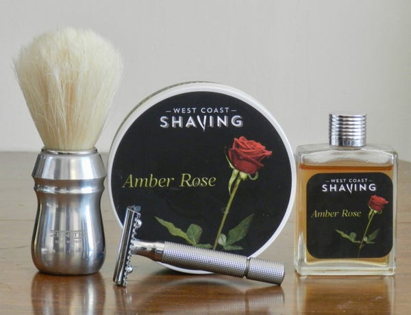 A boar brush with a large bleached knot and aluminum handle stands next to a tub of shaving soap whose labe shows a red rose blossom, just opened, on a black background. "Amber Rose" is printed in gold italics next to the image, and in white at the top is "West Coast Shaving." Next to the soap is a small rectangular glass bottle with a silver cap and the same label. Lying on its side in front is a DE razor with a comb guard and a checkquered handle.