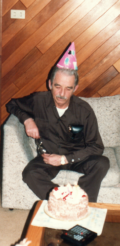 Colour photo of my late Grandfather as he sits in front of a lit birthday cake, which is sitting on an inset glass-panelled coffee table, and decorated in white and red, with white frosting/whipped cream, and red cherries on top.
My Grandfather is wearing a brown shirt and matching pants along with a mostly pink cone-shaped paper party hat.
He's holding his glasses in his hands, vertically above his right leg, as his front pocket is filled with a black leatherette eyeglass case.
The wall behind him is covered in diagonal strips of cedar wood, of which, he had likely installed himself a number of years prior, as he was instrumental in building the home this photo was taken in, which was my aunt's. A unique feature of my aunt's house which can be seen in the upper left corner was the use widespread installation of push button light switches. Never really saw that particular type in houses I visited then.