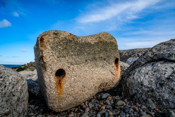 Stone block with two equal distance metal pipes through it. Rust gives impression  of tears out of the pipe 'eyes'. Blue skies above and pebbles and blocks around